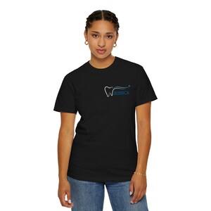 Peut inclure: T-shirt noir avec un petit motif dentaire sur la poitrine. Le motif représente le contour blanc d'une dent avec une ligne bleue incurvée et le nom "JESSICA" en bleu. Le modèle porte un jean bleu.