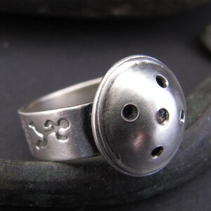May include: A silver ring with a domed top featuring five evenly spaced circular cutouts. The band of the ring is decorated with a stylized design.