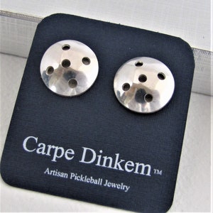 May include: A pair of silver pickleball earrings with a circular design and multiple holes. The earrings are displayed on a black card with the text "Carpe Dinkem Artisan Pickleball Jewelry TM".