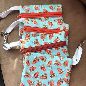 Puede incluir: Varias pequeñas bolsas rectangulares con un estampado floral azul claro con macetas rojas. Cada bolsa tiene una cremallera roja y una correa blanca con un cierre plateado. Las bolsas están dispuestas de forma ligeramente superpuesta.