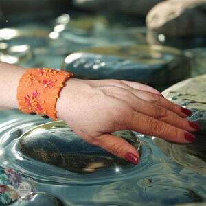 May include: Orange felt bracelet with pink and orange embroidered flowers. The bracelet is on a person's wrist, with their hand resting on the surface of water.