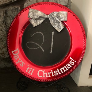 May include: A red charger plate with a black chalkboard center. The chalkboard has the number 21 written on it and the words "Days 'til Christmas!" written in white. A silver glitter bow is on top of the chalkboard.