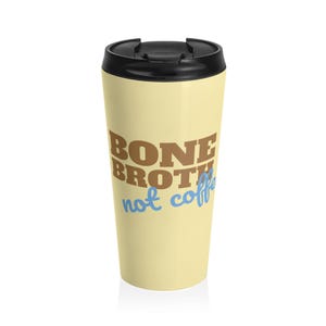 以下が含まれることがあります： 黒い蓋が付いたクリーム色のトラベルマグ。マグには、茶色で「BONE BROTH」、青色で「not coffee」と書かれています。マグは、飲み物を温かくまたは冷たく保つように設計されています。