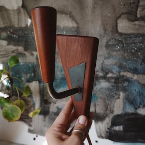 Könnte beinhalten: Eine Wandleuchte mit einer dreieckigen Holzrückplatte und einer dunklen Metalleinlage. Ein gebogener Messingarm ragt aus der Mitte hervor und trägt einen konischen, zylindrischen Holzlampenkopf. Ein dünnes Stromkabel hängt an der Unterseite.