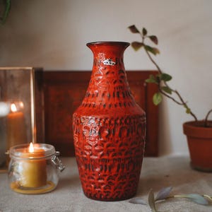 Peut inclure: Un vase en céramique rouge vif avec une surface texturée et ornée de motifs. Le vase a un col étroit et un bord légèrement évasé. Une petite étiquette est visible en haut. Un objet décoratif pour les fleurs ou comme pièce unique.