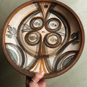 May include: A round ceramic bowl featuring a speckled cream base with hand-painted brown and blue abstract patterns. The design includes four prominent circular motifs arranged in a square formation, accented by dark brown lines and leaf-like shapes.