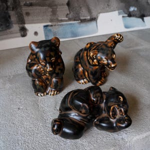 Op de afbeelding: Drie keramische berenbeeldjes met een glanzende afwerking en een gevlekt bruin en zwart kleurpatroon. Eén beer zit rechtop met de poten bij het gezicht, een andere staat met één poot omhoog en de derde ligt op de rug met de poten omhoog.