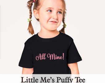 May include: A black t-shirt with the pink text "All Mine!" is worn by a young girl with braided hair. The text "Little Me's Puffy Tee" is displayed below the shirt. The girl is smiling and looking to the side.