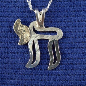 Puede incluir: Un collar con un colgante de plata y un dije en forma de letra hebrea 'Chai' de color dorado. El dije está finamente diseñado con una superficie texturizada.
