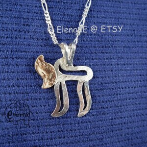 Puede incluir: Un collar de plata con un colgante en forma de la letra hebrea 'Chai' con un acabado texturizado. El colgante tiene un pequeño acento dorado en el lateral.