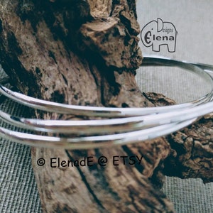 May include: Three silver bangles stacked on top of each other on a piece of driftwood. The bangles are smooth and shiny. The text "Designs by Elena" is visible in the top right corner of the image.