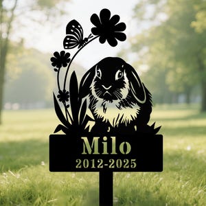May include: Black metal garden stake featuring a rabbit silhouette with a butterfly and flowers. The stake includes the name "Milo" and the years 2012-2025. The design is set against a blurred green background.