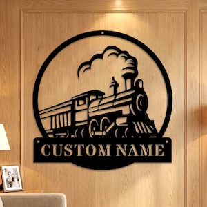 Op de afbeelding: Een rond wandbord van zwart metaal met een silhouet van een stoomlocomotief die rook uitstoot. Onder de trein bevindt zich een rechthoekige banner met de tekst CUSTOM NAME in vetgedrukte hoofdletters en twee gaten voor montage.