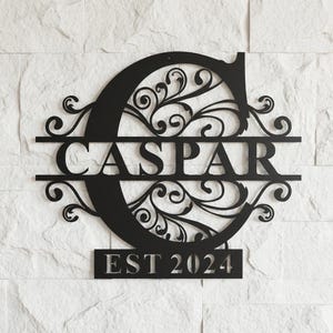 May include: Black metal wall art with the name "CASPAR" displayed within a decorative "C" initial. The design incorporates scrolling patterns and the text "EST 2024" at the base, ideal for interior decoration.