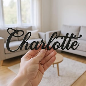 May include: A black cursive name cutout reading "Charlotte". The name is displayed against a living room backdrop with a sofa, rug, and small table. The name is in a stylish font.