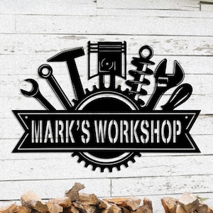 May include: Black metal workshop sign featuring tools like a hammer, wrench, and piston. The sign has the text "MARK'S WORKSHOP" in a banner across the center, with a gear design below.