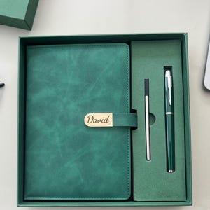 May include: A green leather journal and pen set in a gift box. The journal has a gold clasp with the name "David" engraved. The set includes a pen and a stylus, all in matching green. The box is also green.