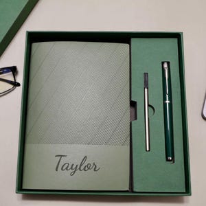 Può includere: Un set regalo verde contenente un taccuino, una penna e una ricarica. Il taccuino ha il nome "Taylor" stampato sopra. Il set è presentato in una scatola verde.