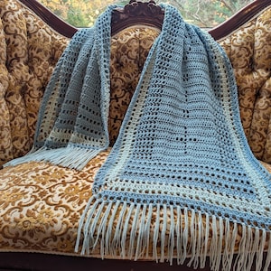 Puede incluir: Bufanda de ganchillo azul claro con flecos, colocada sobre un sofá vintage con estampado floral. El patrón de ganchillo de la bufanda incluye calados y un borde azul más claro. Los flecos son de color azul claro a juego.