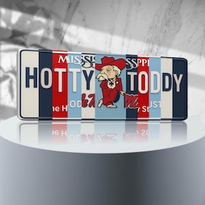 May include: A rectangular metal sign with the words "HOTTY TODDY" in white block letters. The sign has a colorful design with red, blue, and white stripes and a cartoon illustration of a man in a red suit and cowboy hat. The word "Mississippi" is at the top.
