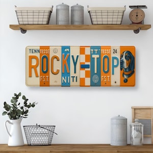 K&ouml;nnte beinhalten: Rechteckiges Metallschild mit Vintage-Kennzeichen-Design. Das Schild zeigt die Worte "ROCKY TOP" in Orange und Blau, mit einer Grafik eines Jagdhundes auf der rechten Seite. Das Schild ist cremefarben mit orangefarbenen und blauen Akzenten.