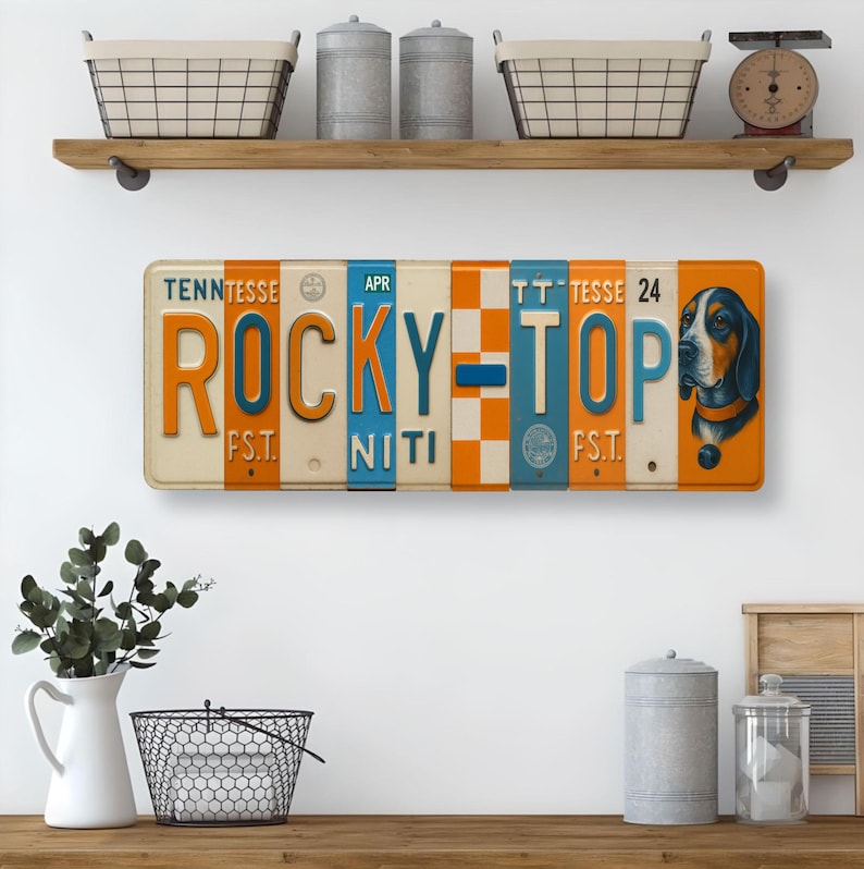 K&ouml;nnte beinhalten: Rechteckiges Metallschild mit Vintage-Kennzeichen-Design. Das Schild zeigt die Worte "ROCKY TOP" in Orange und Blau, mit einer Grafik eines Jagdhundes auf der rechten Seite. Das Schild ist cremefarben mit orangefarbenen und blauen Akzenten.