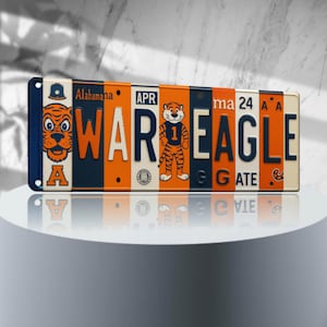 May include: A vintage-style metal license plate with the words "WAR EAGLE" in orange and blue letters. The plate has a cartoon tiger mascot wearing a jersey with the number "1" on it. The plate also has the words "Alabama" and "GATE" on it.