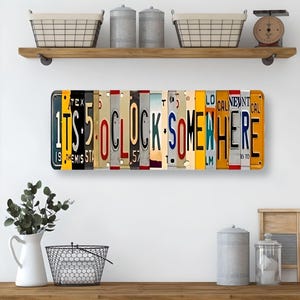 May include: A metal sign made of repurposed license plates spelling out "It's 5 o'clock somewhere". The sign is hanging on a shelf above a kitchen counter.