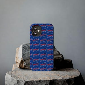 Puede incluir: Una funda de teléfono azul con un patrón repetido del logotipo de los Buffalo Bills en blanco, rojo y azul. La funda se muestra sobre una superficie de piedra texturizada sobre un fondo gris. La funda del teléfono tiene un módulo de cámara.