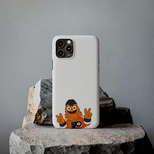 Op de afbeelding: Een witte telefoonhoes met een cartoonmascotte met oranje vacht, een zwarte hoed en het logo van de Philadelphia Flyers. De mascotte maakt het vredesteken. De telefoonhoes ligt op een grijs stenen oppervlak.