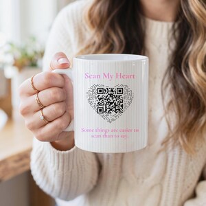 May include: A white ceramic mug with a subtle vertical pinstripe pattern. The front features the pink text Scan My Heart above a black and white QR code shaped like a heart, with the pink text Some things are easier to scan than to say below.