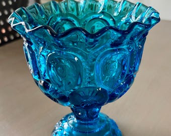 May include: A blue glass vase with a scalloped rim and a textured, star-shaped pattern. The vase has a pedestal base.