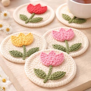 May include: Crocheted coasters featuring tulip designs in red, yellow, and pink. The coasters have a cream-colored base with green stems and leaves. A cup of tea sits on one of the coasters, and daisies are scattered around.