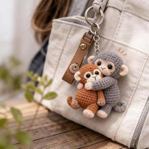 Op de afbeelding: Een handgemaakte gehaakte sleutelhanger met twee knuffelende apen. Eén aap is grijs, de andere bruin. De sleutelhanger heeft een bruin leren bandje met een smiley en een zilveren metalen ring.