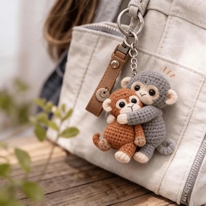 May include: A handmade crochet keychain featuring two hugging monkeys. One monkey is brown, the other is gray. The keychain has a brown leather strap with a smiley face charm and a silver keyring. The background includes a white bag and a wooden surface.