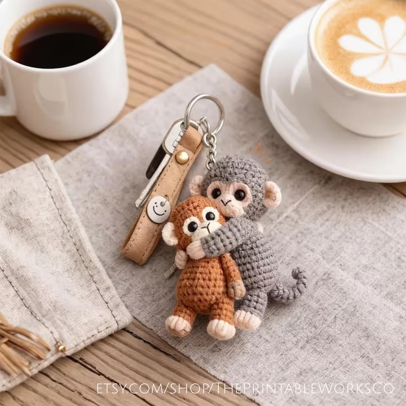 May include: A handmade crochet keychain featuring two monkey figures, one hugging the other. The monkeys are in shades of brown and gray, with white accents. The keychain includes a leather strap with keys and a metal ring. The Etsy shop name is visible.