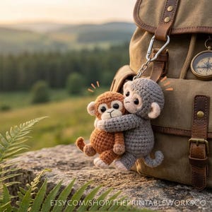 May include: A crocheted keychain featuring two hugging monkeys. One monkey is brown, the other is gray. The keychain is attached to a tan backpack with leather straps and a compass. The background is a blurred view of a green landscape.