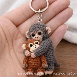 May include: A handmade crochet keychain featuring two small monkeys. One gray monkey hugs a smaller brown monkey. The keychain has a silver ring. The Etsy shop URL is visible at the bottom of the image.