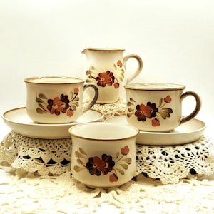 May include: A vintage cream ceramic tea set with floral patterns. The set includes two teacups with saucers, a creamer jug, and a small bowl. Each piece has a brown rim and a design of red and brown flowers with green leaves.