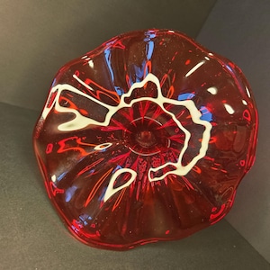 May include: A red glass bowl with a wavy rim and a white, abstract design in the center. The bowl has a glossy finish and is likely handmade. The bowl's diameter is approximately 20 cm.