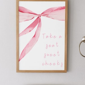 Könnte beinhalten: Gerahmter Druck mit einem rosa Aquarell-Schleifendesign und dem Text "Take a seat sweet cheeks". Der Druck befindet sich in einem hellbraunen Holzrahmen und hängt über einer weißen Toilette.