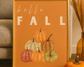 May include: A framed art print with the words "hello FALL" in white on an orange background. The print features a cluster of watercolor pumpkins in shades of orange, green, and brown. A real pumpkin sits nearby.