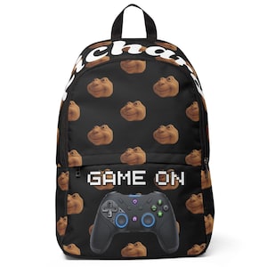 Op de afbeelding: Zwarte rugzak met een patroon van bruine cartoongezichten. De rugzak heeft een zwart handvat en een zwarte controller-afbeelding met de woorden "GAME ON" in het wit. Het woord "cha" staat in het wit bovenaan.
