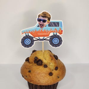 May include: A chocolate chip muffin topped with a decorative cupcake pick. The pick features a cartoon monster truck design with a child's face wearing sunglasses. The truck is light blue with orange flames and blue wheels.