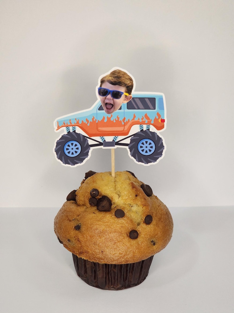 May include: A chocolate chip muffin topped with a decorative cupcake pick. The pick features a cartoon monster truck design with a child's face wearing sunglasses. The truck is light blue with orange flames and blue wheels.