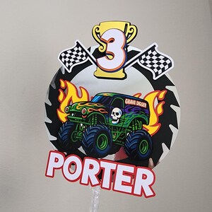 Pode incluir: Topper de bolo colorido com um design de monster truck, o texto "Grave Digger" e o nome "Porter". Inclui o n&uacute;mero "3" em um trof&eacute;u dourado, bandeiras quadriculadas e chamas.