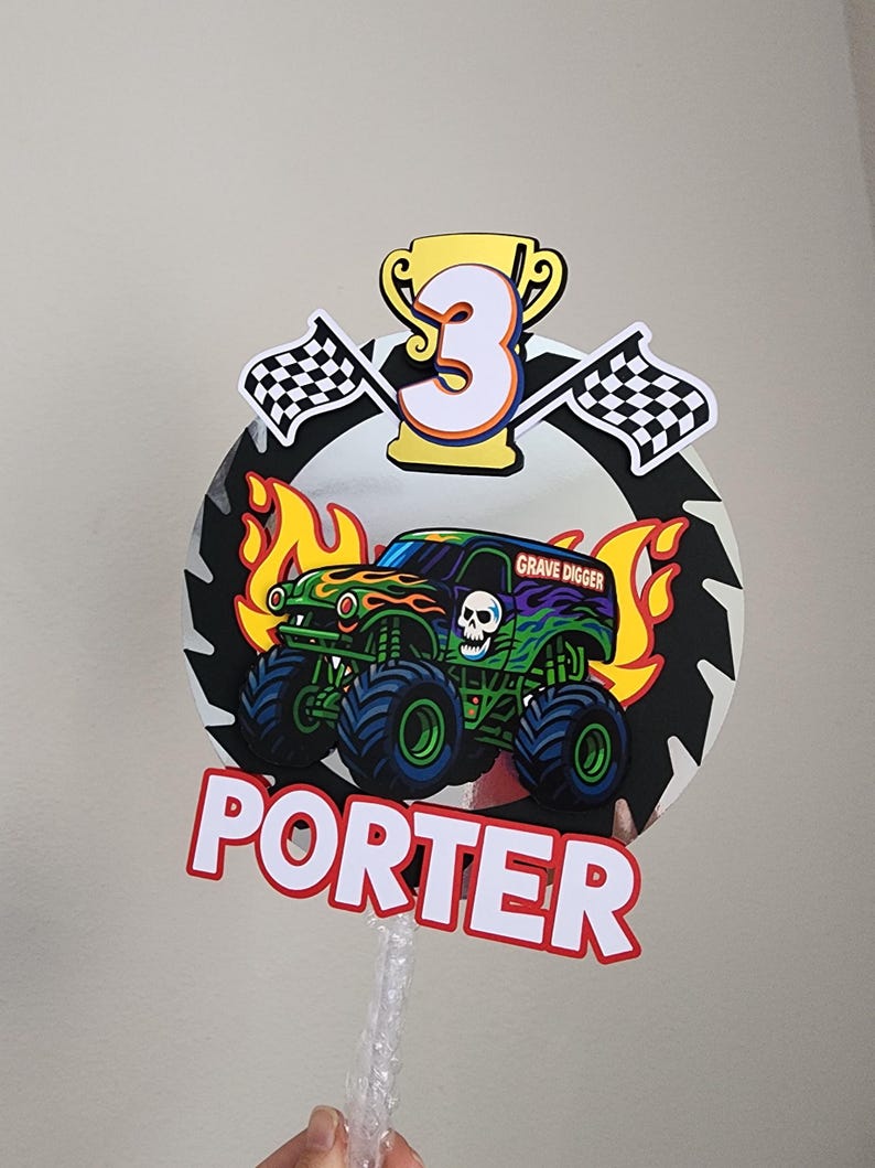 Pode incluir: Topper de bolo colorido com um design de monster truck, o texto "Grave Digger" e o nome "Porter". Inclui o n&uacute;mero "3" em um trof&eacute;u dourado, bandeiras quadriculadas e chamas.