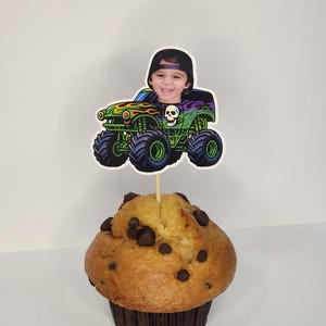 May include: A cupcake topped with a monster truck-themed decoration. The decoration features a child's face in a monster truck design with green, black, and red accents. The cupcake is a muffin with chocolate chips.