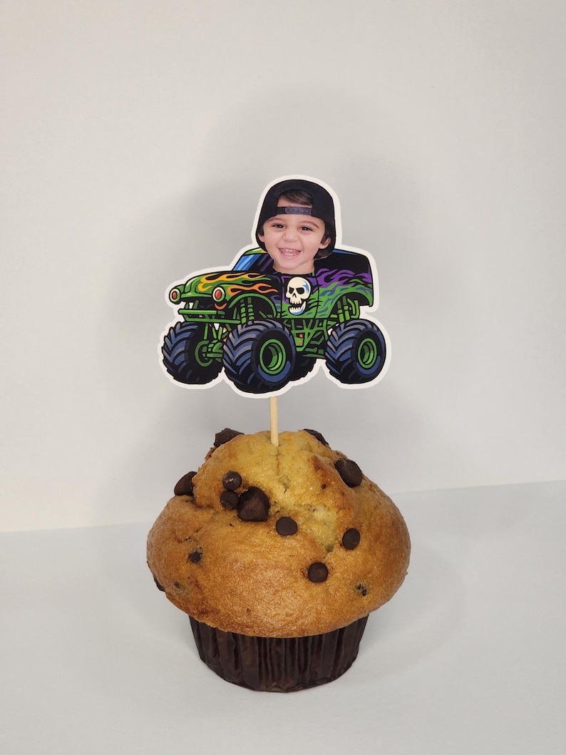 May include: A cupcake topped with a monster truck-themed decoration. The decoration features a child's face in a monster truck design with green, black, and red accents. The cupcake is a muffin with chocolate chips.