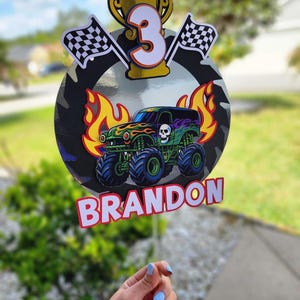 Pode incluir: Decora&ccedil;&atilde;o de anivers&aacute;rio colorida com um monster truck, chamas, uma caveira e o n&uacute;mero 3. Inclui bandeiras quadriculadas, um trof&eacute;u e o nome "BRANDON". O fundo apresenta um padr&atilde;o de camuflagem, ideal para uma festa.